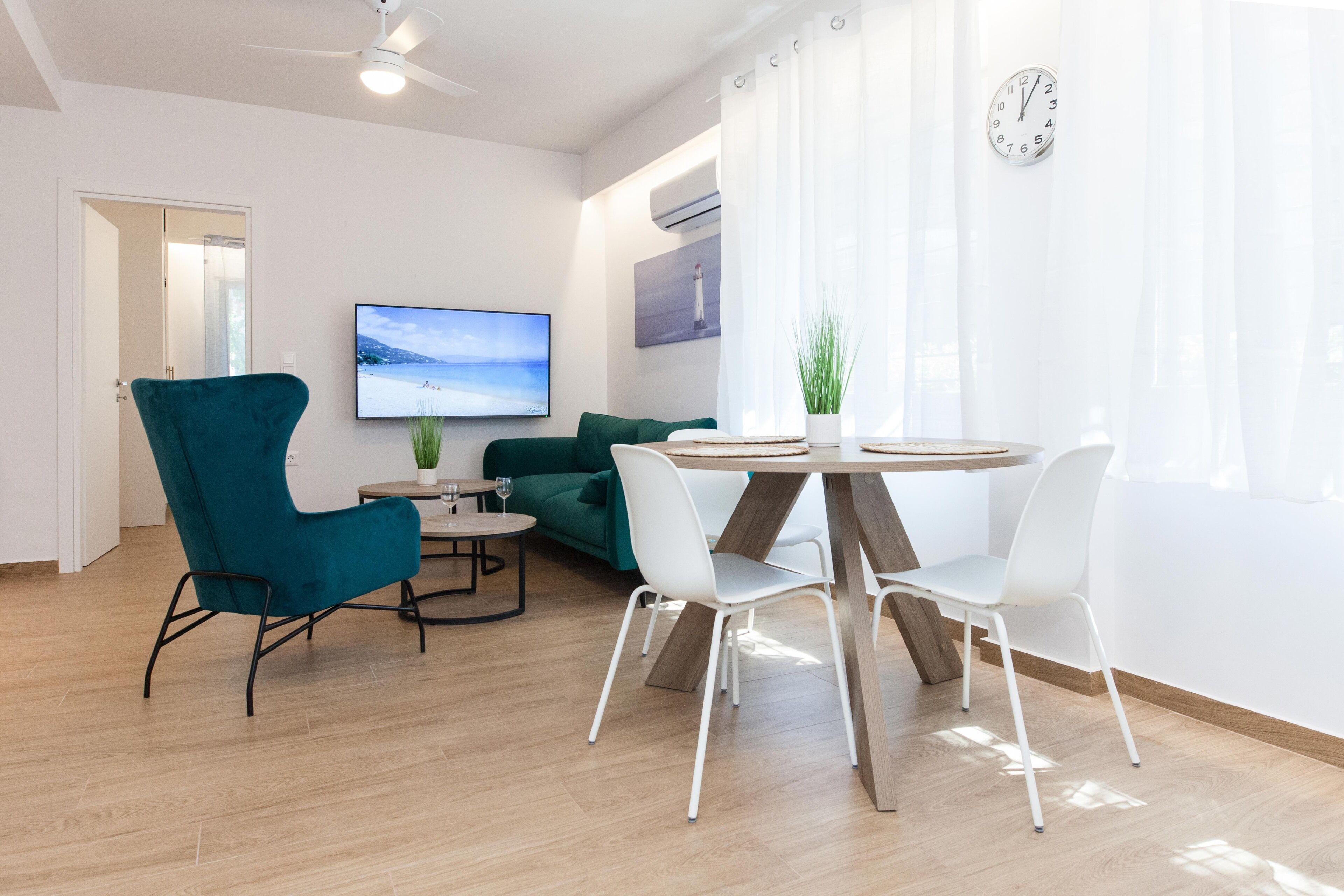 Een moderne woonkamer met een houten vloer, een blauwgroene fauteuil, witte stoelen, een ronde tafel, een televisie en een klok aan de muur.
