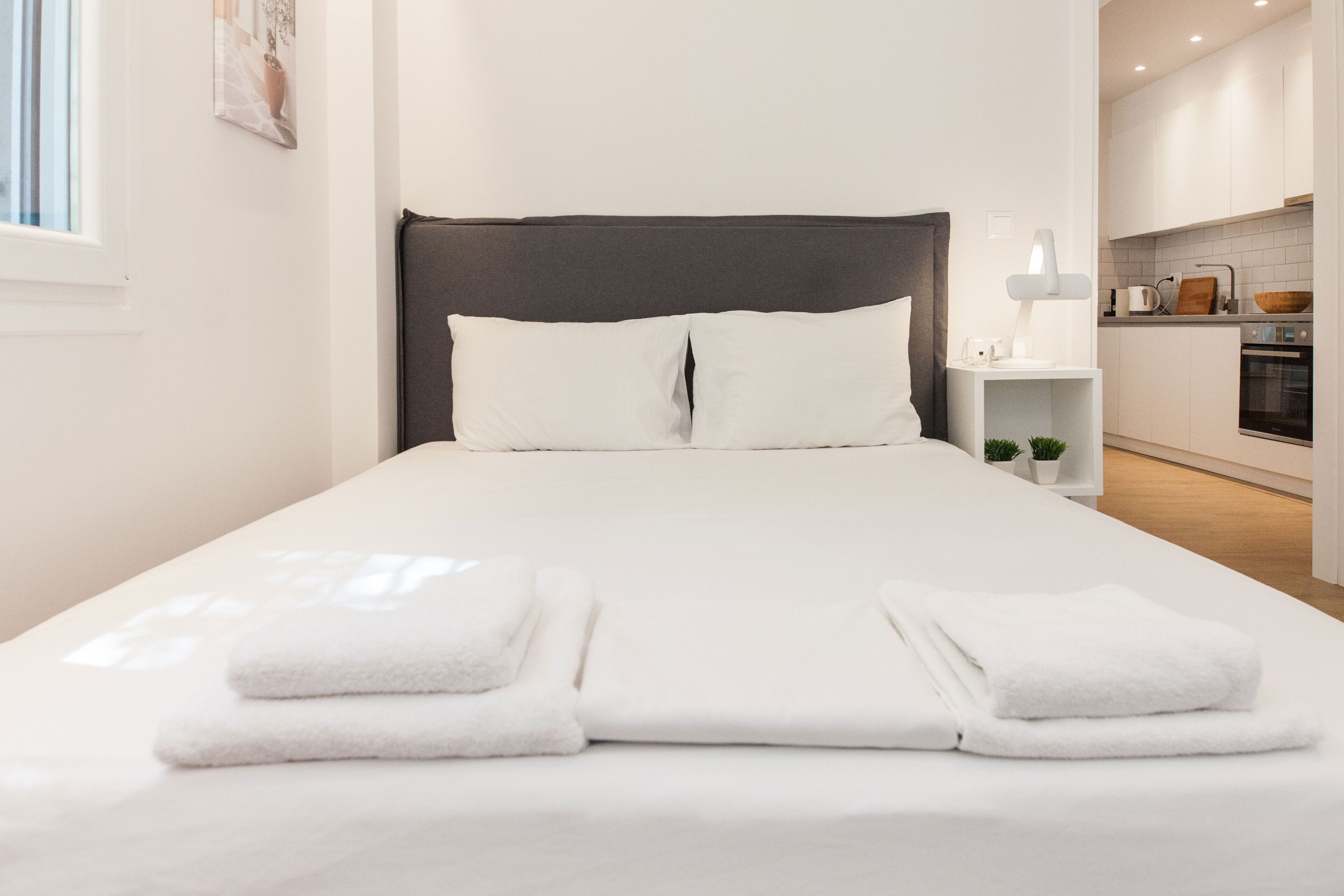 Una cama bien hecha con sábanas blancas y dos toallas dobladas, una mesita de noche con lámpara, y vistas hacia una cocina moderna con armarios blancos y encimera de madera.