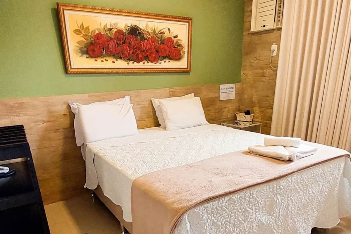Habitación de hotel con cama, mesita de noche, un cuadro de flores y una ventana con cortinas.