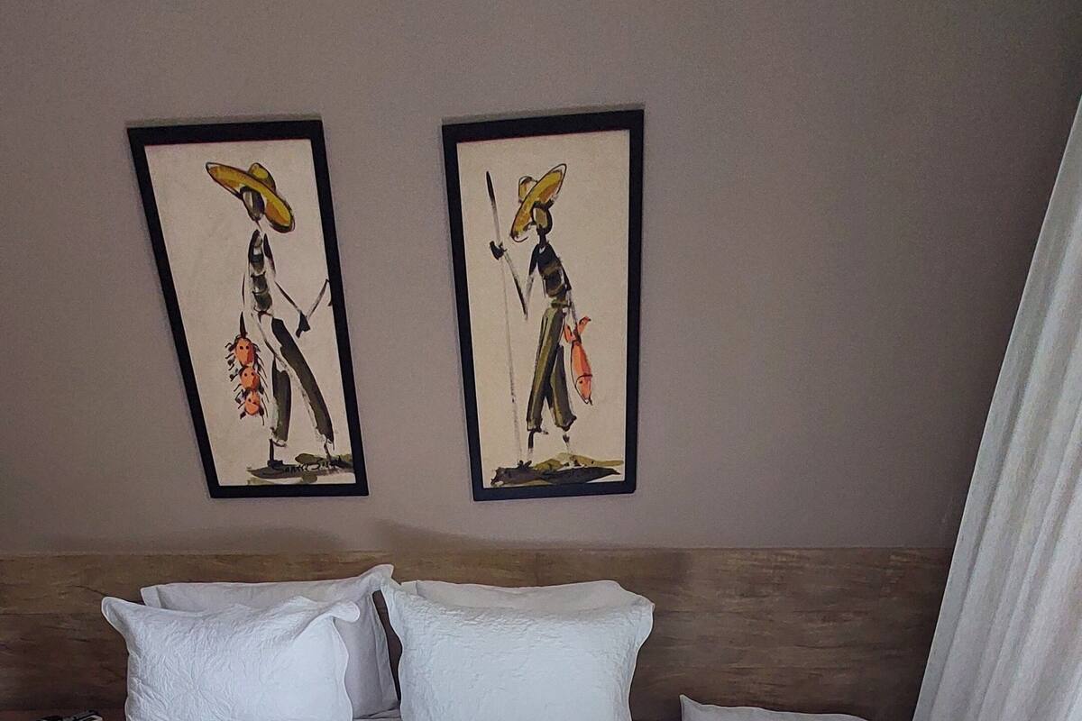 Una cama doble con una colcha azul y blanca, dos almohadas blancas y una manta roja. Encima de la cama, hay dos obras de arte enmarcadas en la pared.
