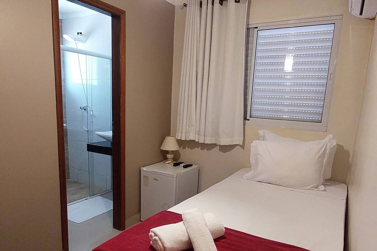 Un dormitorio con cama, mesita de noche, lámpara, ventana con persianas y puerta que da a un baño.