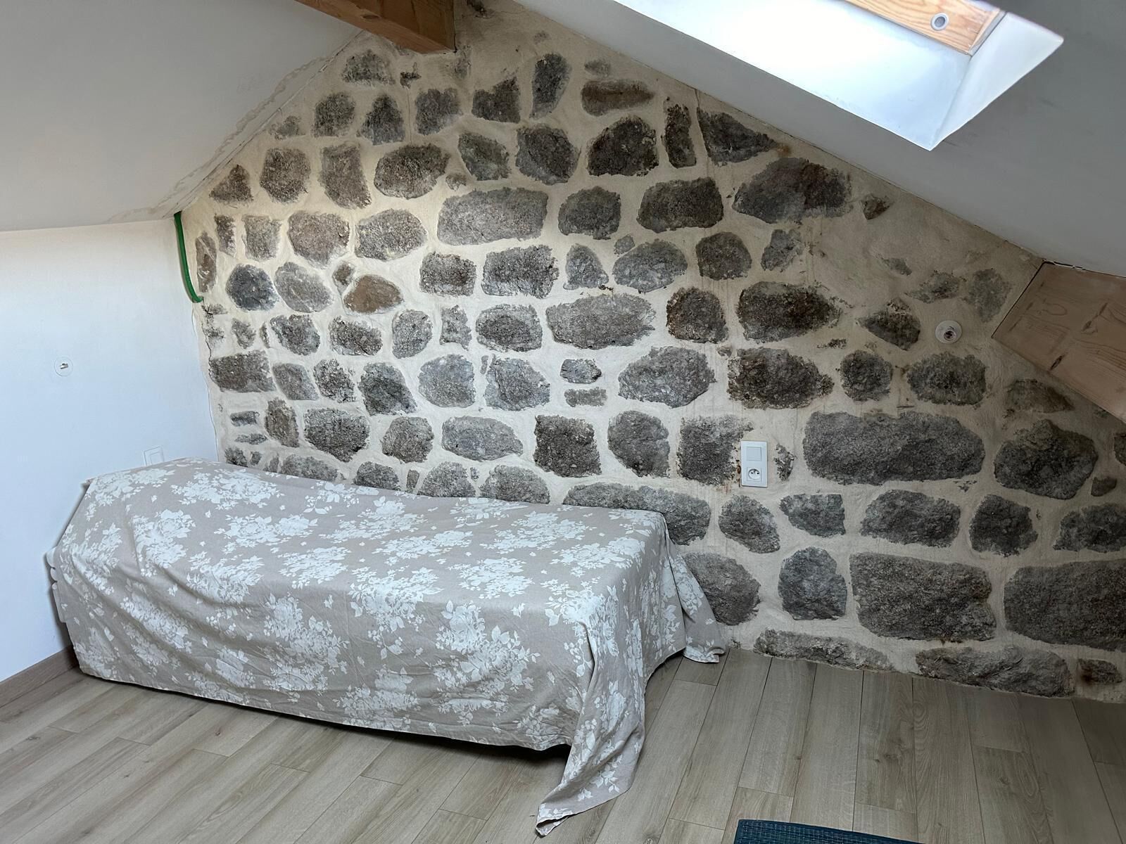 Ein Zimmer mit einer schrägen Steinwand, einem Bett mit floralem Überwurf und einem Oberlicht.
