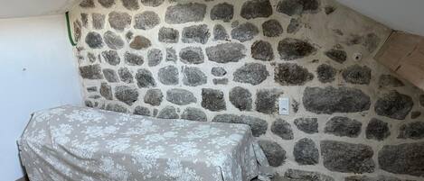 Ein Zimmer mit einer schrägen Steinwand, einem Bett mit floralem Überwurf und einem Oberlicht.