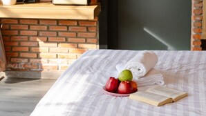 A bed with a white blanket, a book, and a bowl of fruit. There is a potted plant and a radio on a shelf. - Scent Bungalow (Xuan Huong - Da Lat)