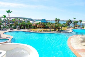 A resort pool with lounge chairs, palm trees, and a view of the ocean and a bridge. - Caribe Penthouse B106 (Orange Beach)