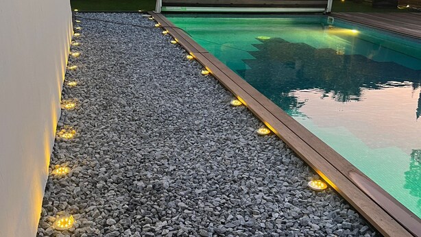 A swimming pool with a pebble-lined deck, wooden fence, and ambient lighting. - Haus in Köln Thielenbruch (Köln)