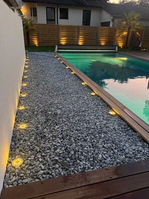 Ein Schwimmbecken mit Kiesdeck, Holzzaun und stimmungsvoller Beleuchtung.