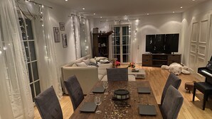 A modern living room with a wooden dining table, grey sofas, and a large TV. - Haus in Köln Thielenbruch (Köln)