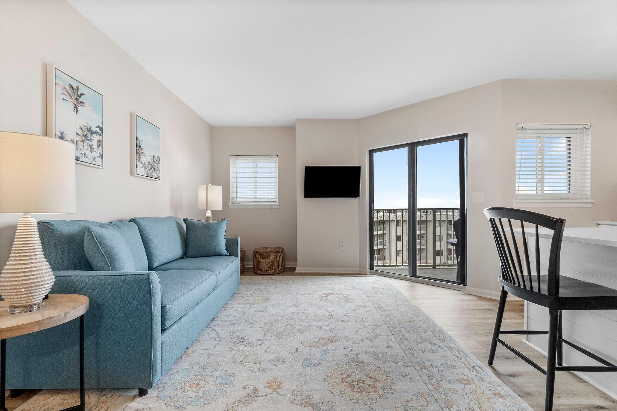 A modern living room with a blue sofa, a round table with a lamp, a flat-screen TV, and a balcony with a view.
