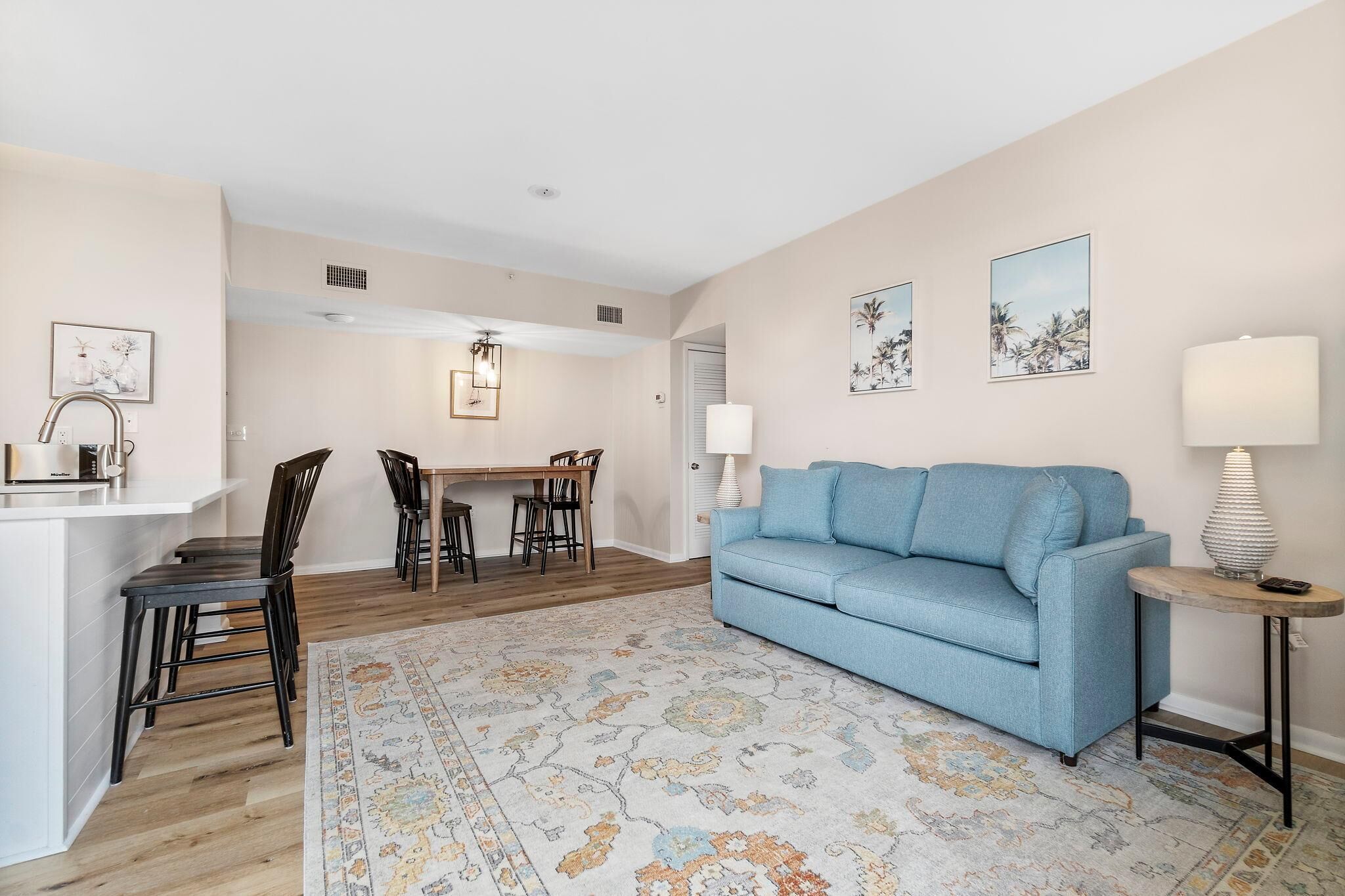 A living room with a blue sofa, a dining table with chairs, a kitchen area, and a patterned rug.