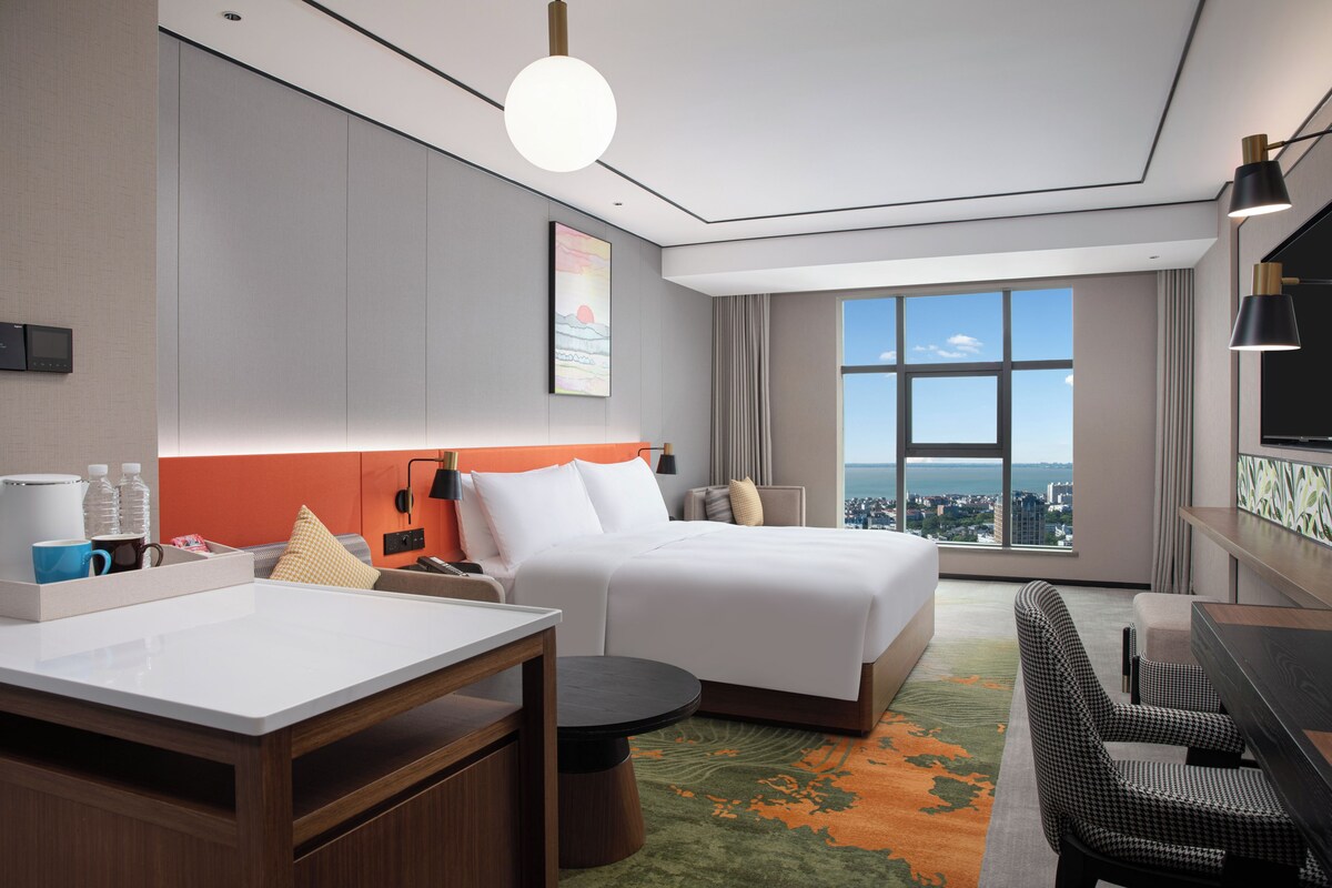 Ein Hotelzimmer mit einem großen Bett, einem Schreibtisch, einem Sessel, einer Kaffeemaschine und Blick auf die Stadt.
