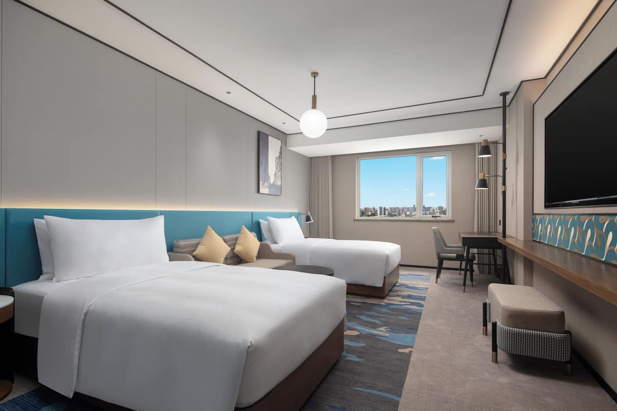 Een hotelkamer met twee bedden, een bureau, een stoel, een televisie en een raam met uitzicht op de stad.