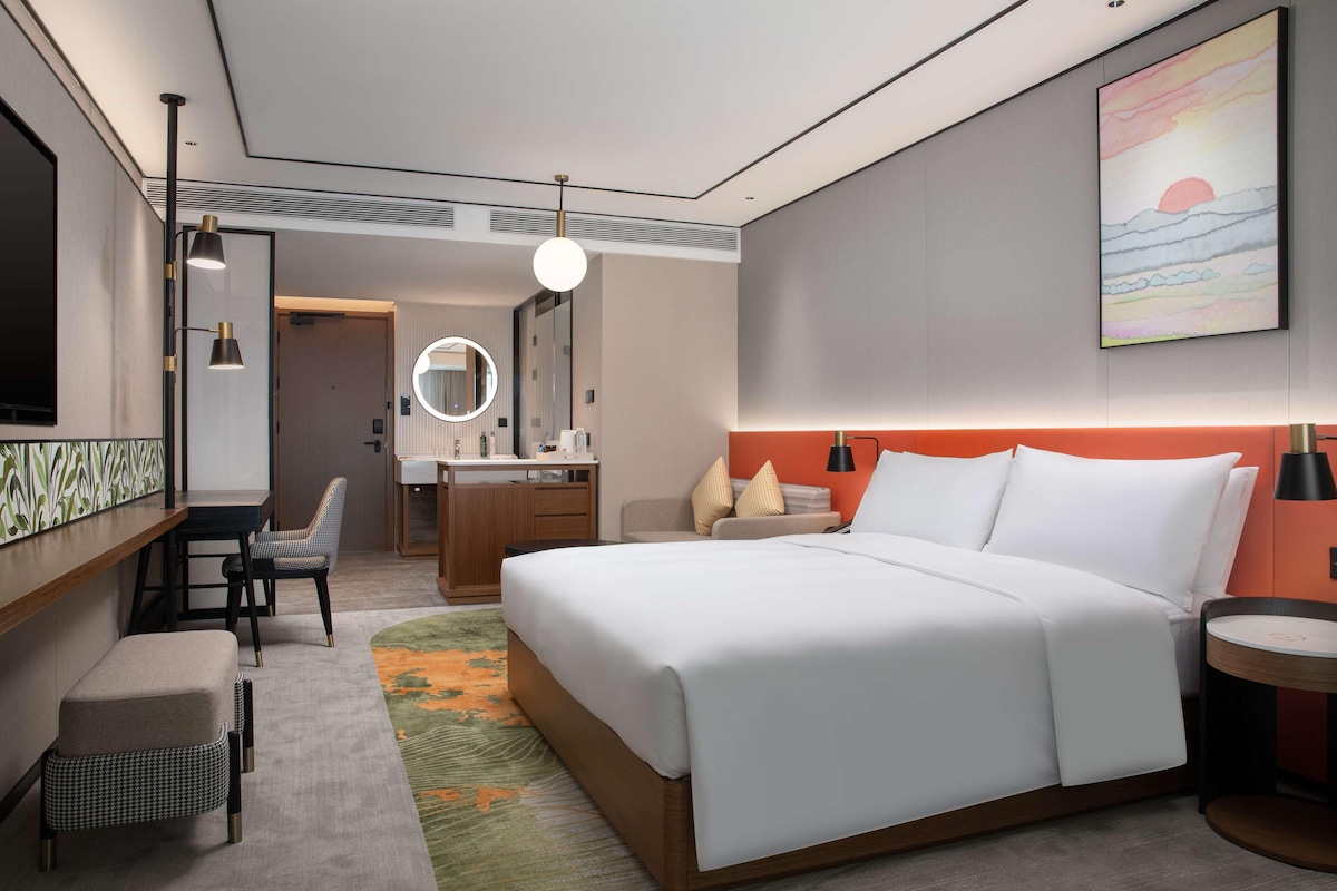 Une chambre d’hôtel moderne avec un grand lit, un bureau, une chaise, une coiffeuse avec un miroir et un tableau au mur.