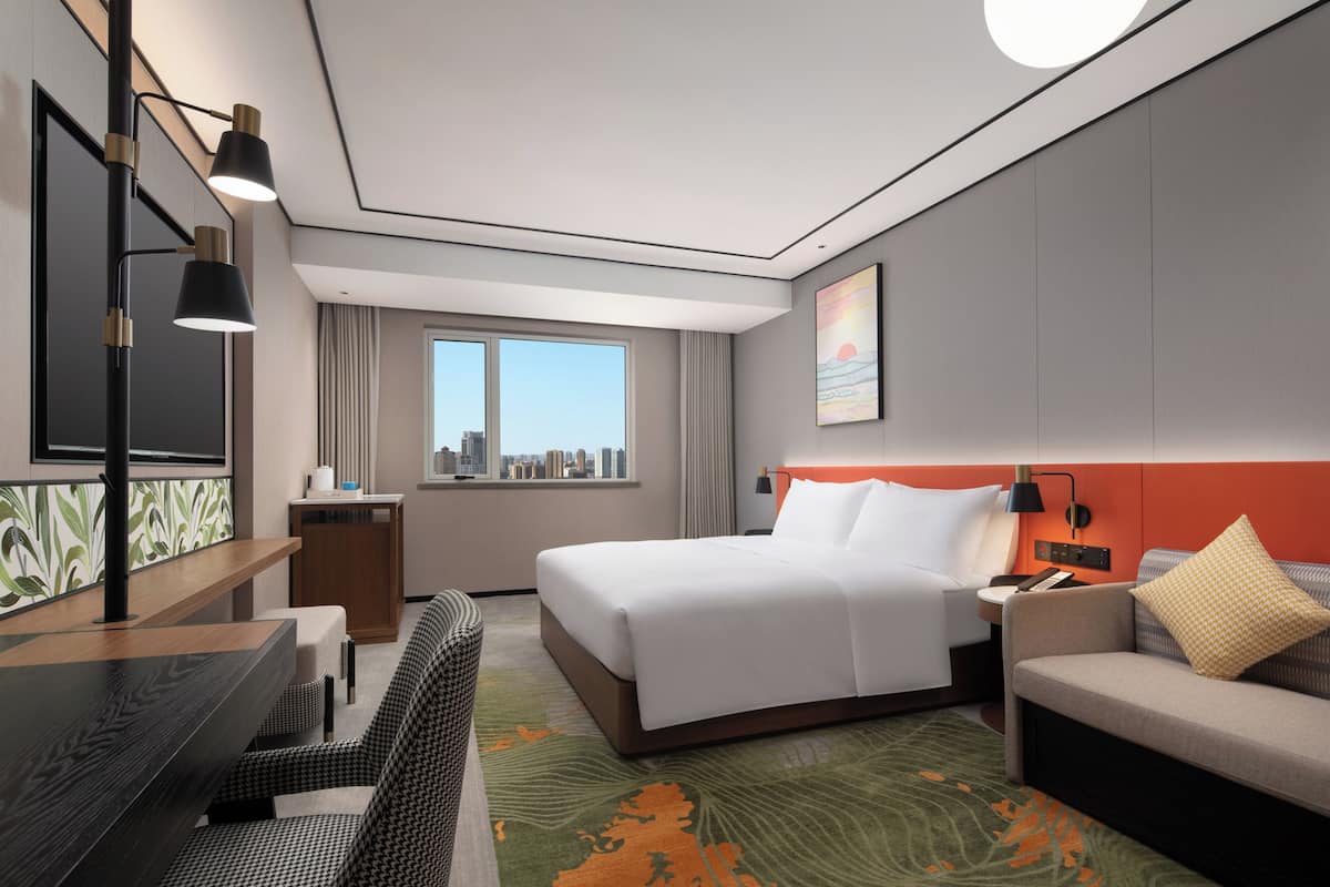 Een hotelkamer met een bed, een bureau, een bank, uitzicht op de stad en een schilderij aan de muur.