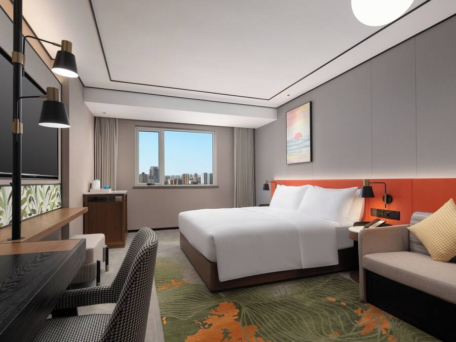 A hotel room with a bed, a desk, a sofa, a city view, and a painting on the wall.