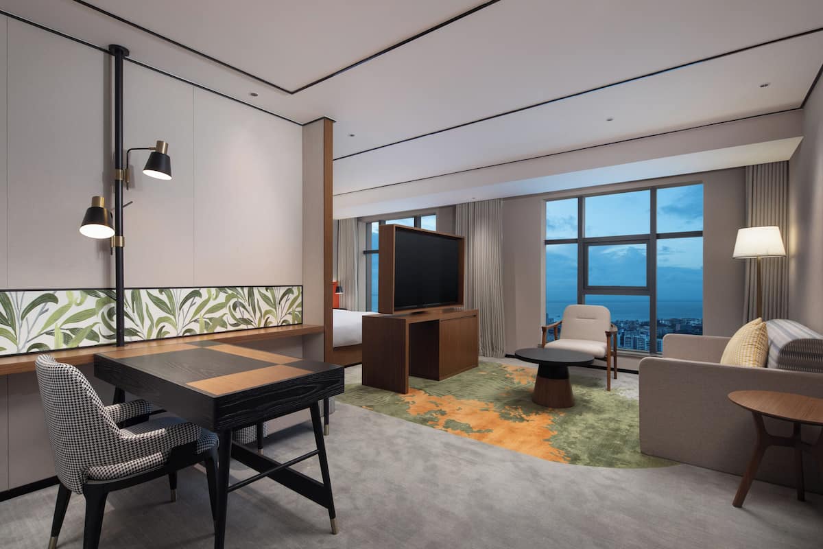 Een moderne hotelkamer met een groot raam, een flatscreen-tv, een zithoek met een tafel en stoelen, en een bank.