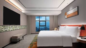 A modern hotel room with a large bed, a flat-screen TV, a city view, and a small seating area.
