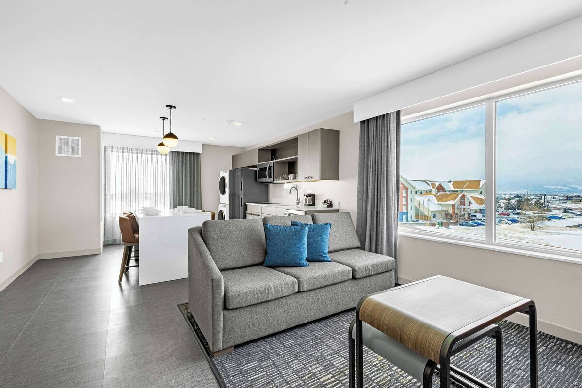 A modern living room with a grey sofa, a small table, and a view of a cityscape through large windows.