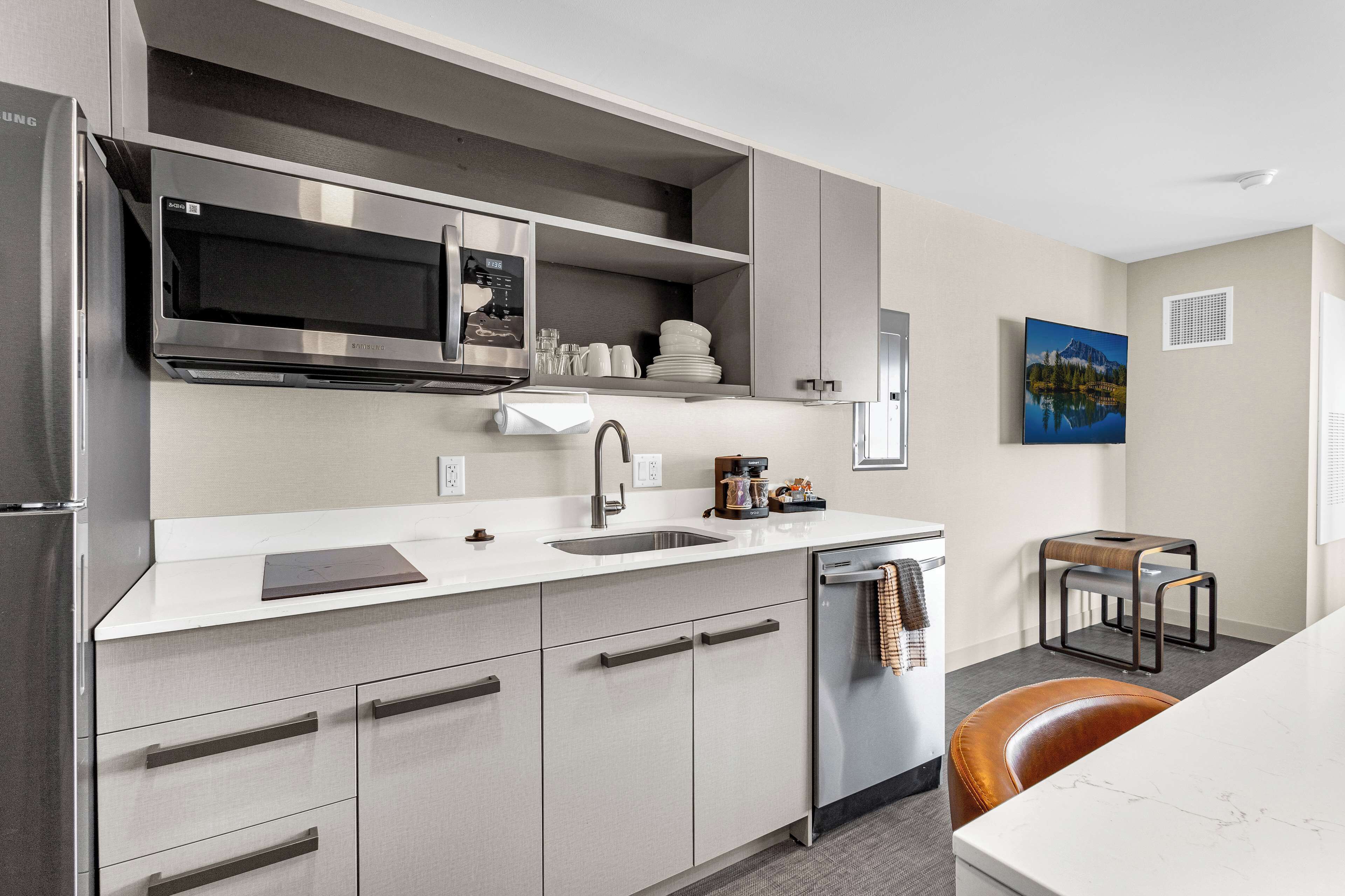 A modern kitchen with stainless steel appliances, a built-in microwave, and a dishwasher. There is a small island with a sink and a towel hanging on the door. A painting is visible on the wall, and a chair is placed near the dining area.