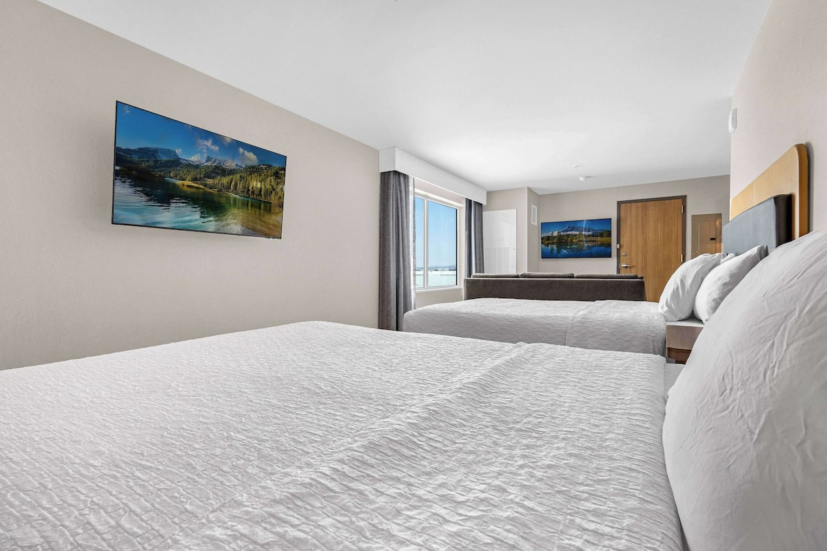 A hotel room with two beds, a large window with a view of mountains, and a painting of a lake.
