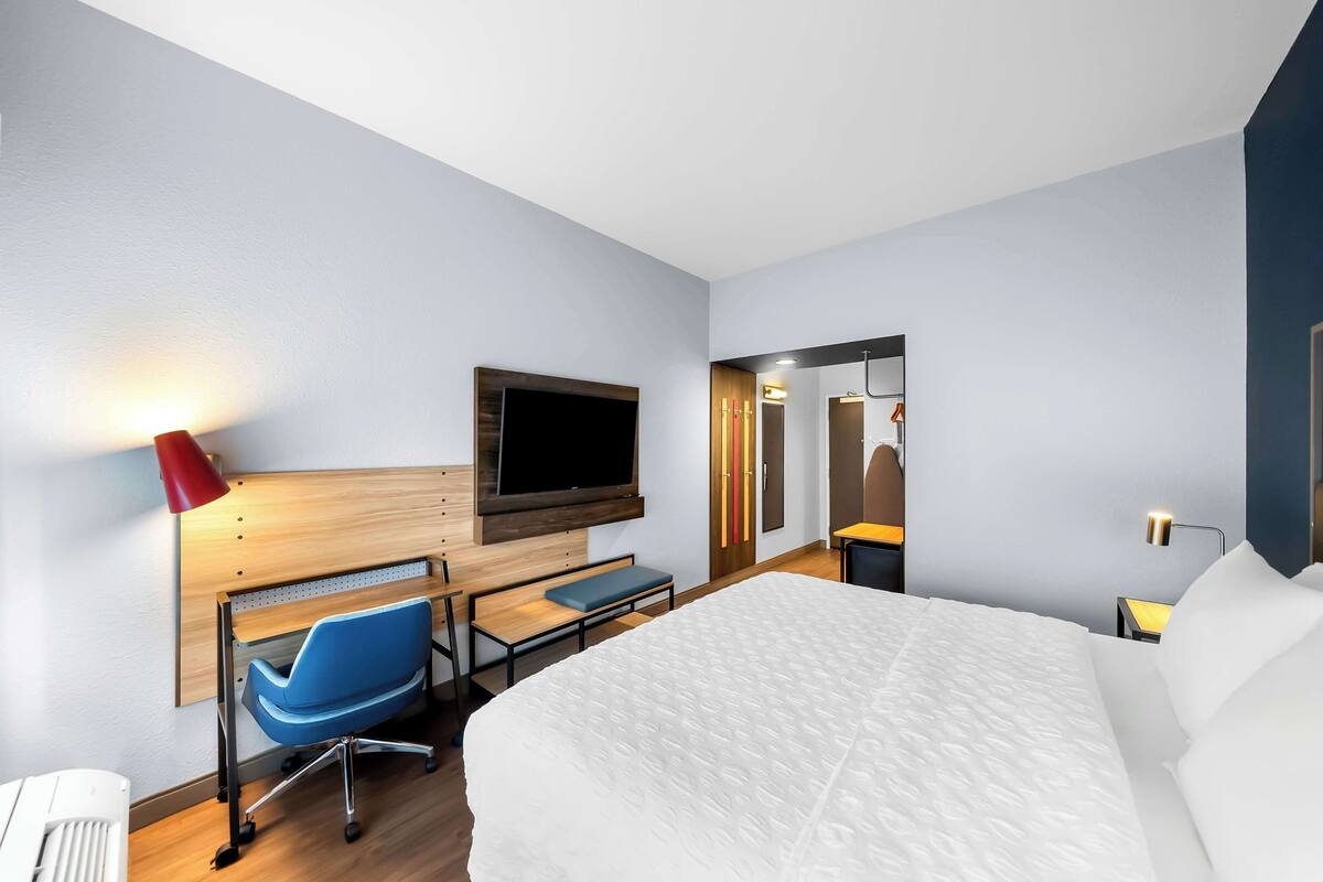 A modern hotel room with a large bed, a desk with a chair, a television mounted on the wall, and a mirror.