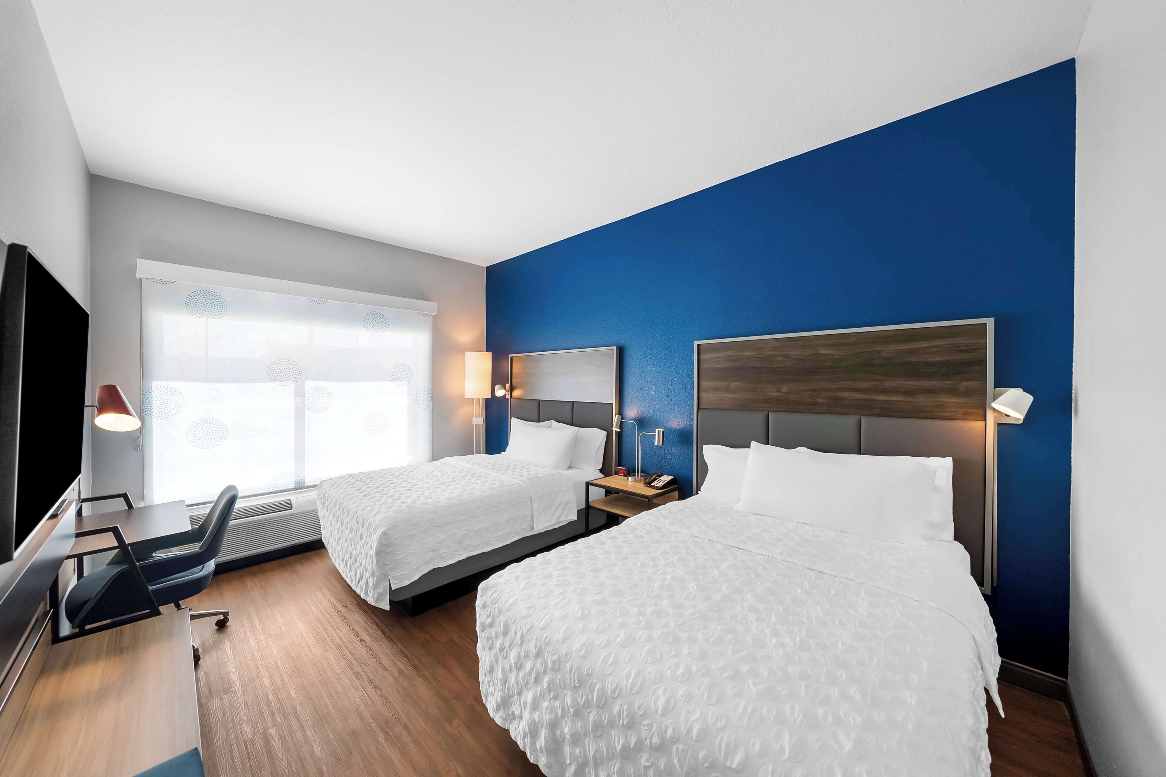 A hotel room with two beds, a desk, a TV, and a window with blinds.