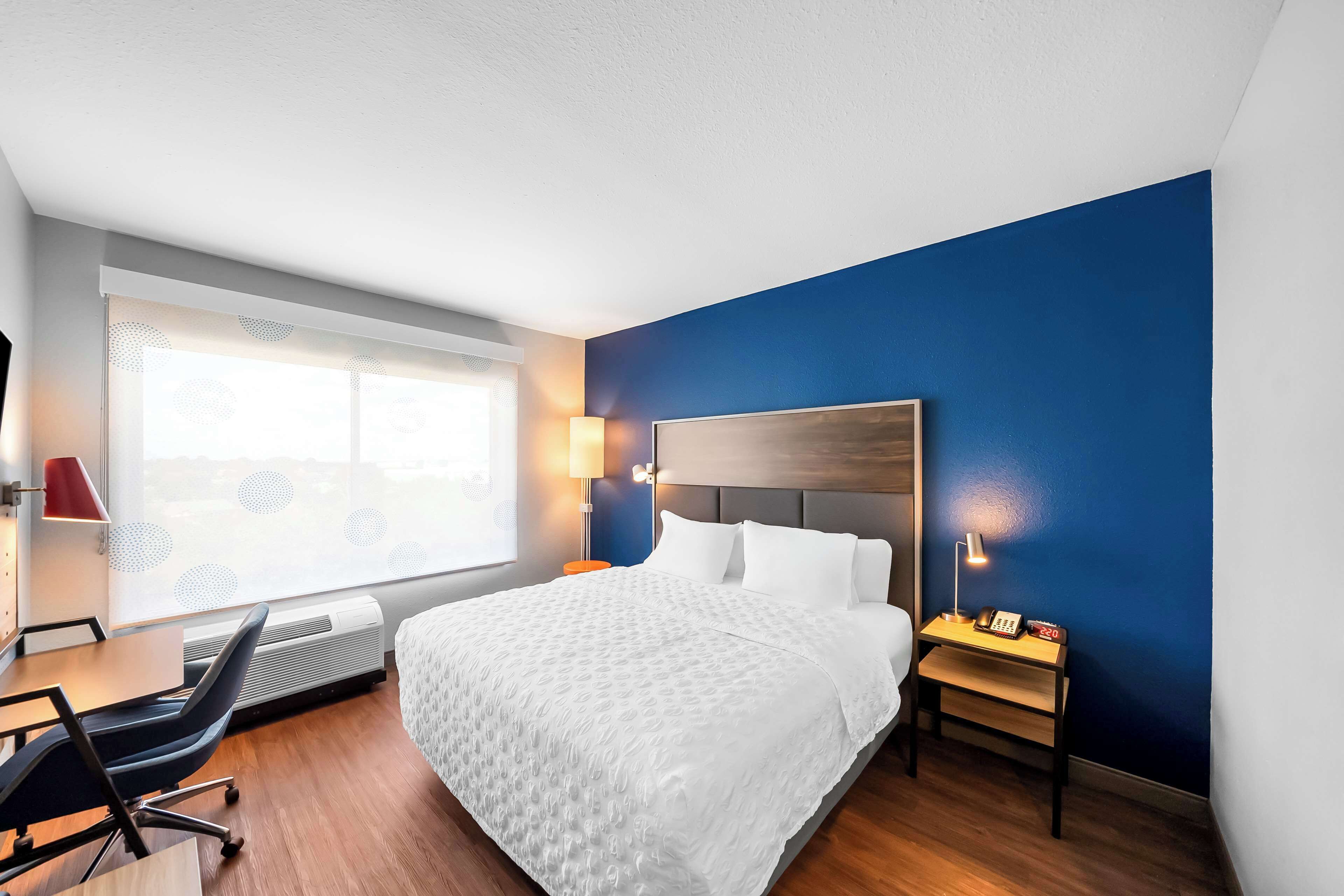 Habitación de hotel con una cama grande, un escritorio con silla, una ventana con persianas y una mesita de noche con lámpara y teléfono.