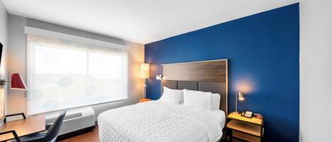 Habitación de hotel con una cama grande, un escritorio con silla, una ventana con persianas y una mesita de noche con lámpara y teléfono.