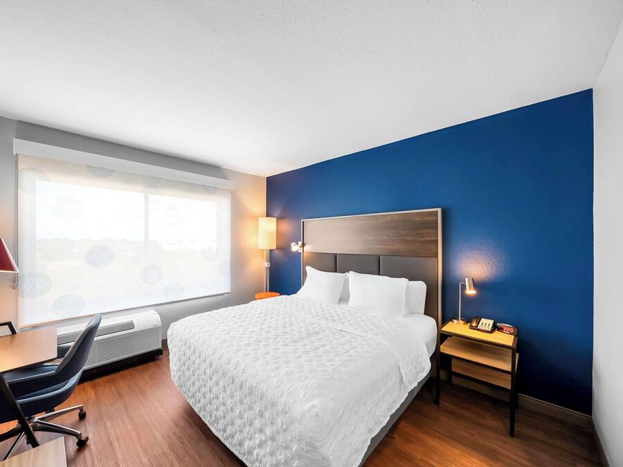 Habitación de hotel con una cama grande, un escritorio con silla, una ventana con persianas y una mesita de noche con lámpara y teléfono.