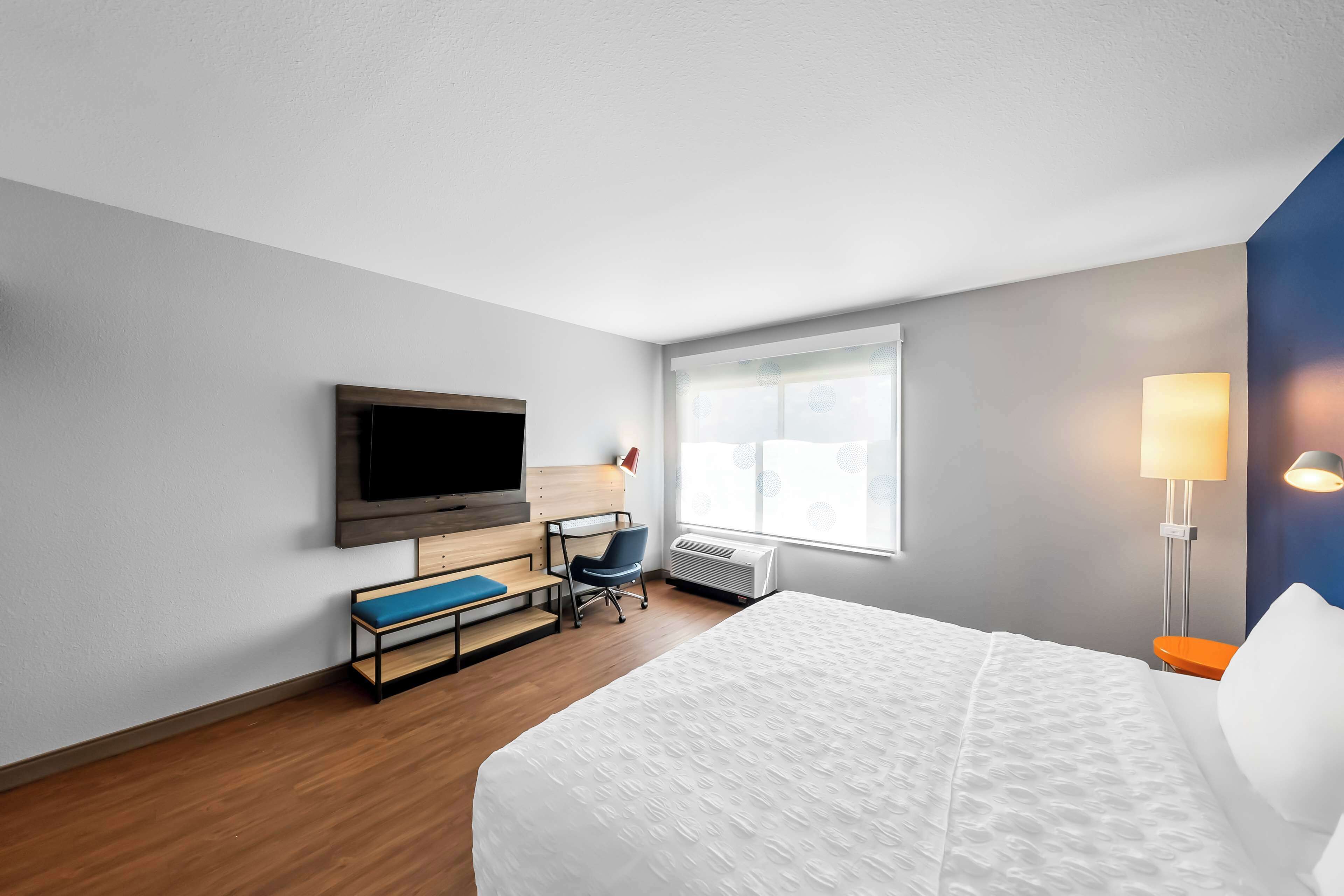 A hotel room with a bed, a desk, a chair, a television, a window with blinds, and a lamp.