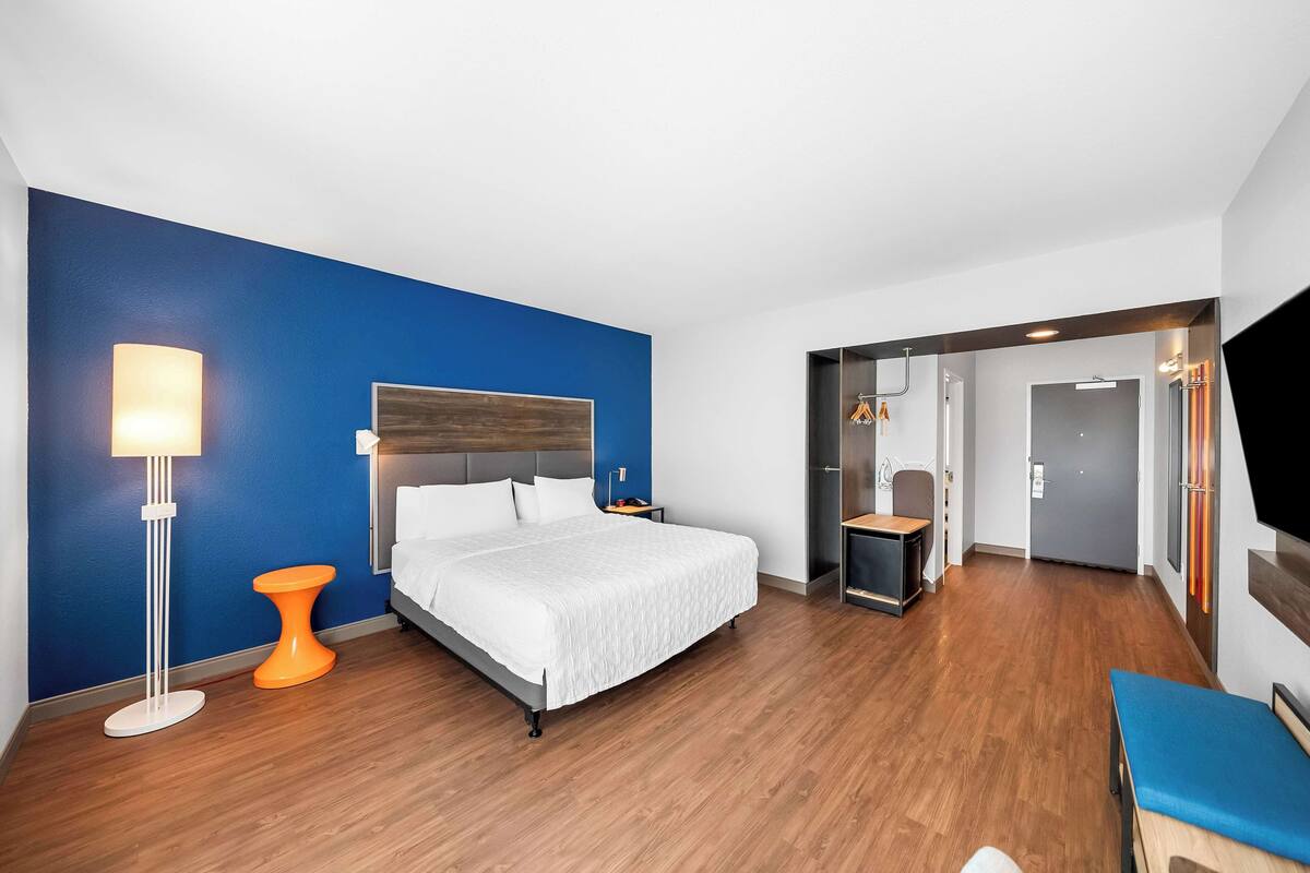A modern hotel room with a large bed, a bedside table, a floor lamp, and a TV mounted on the wall.