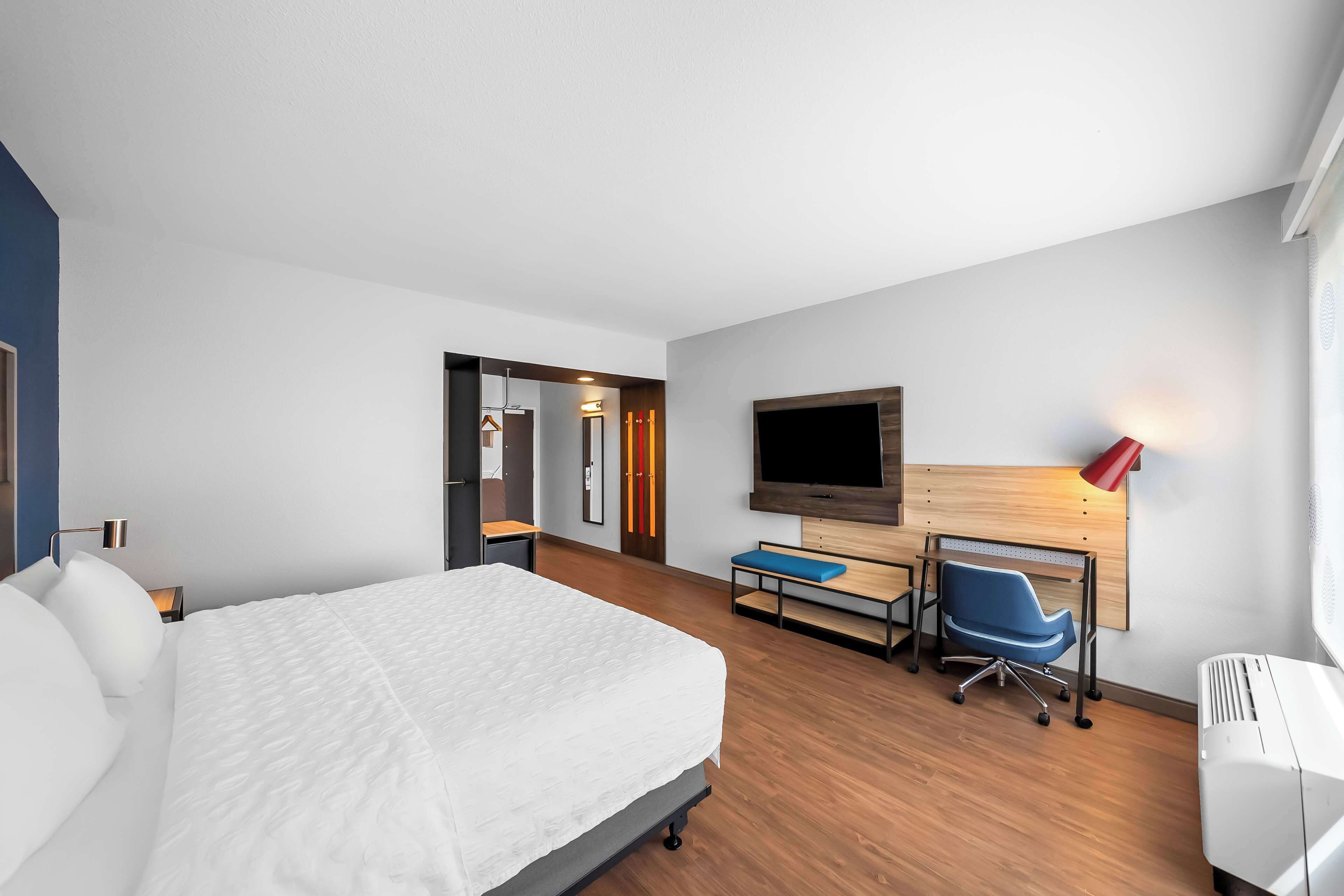 Quarto de hotel moderno com uma cama grande, uma escrivaninha com cadeira, uma TV e um abajur vermelho.