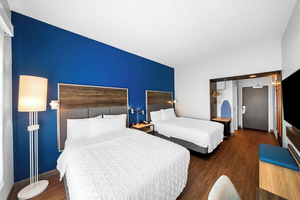 A hotel room with two beds, a desk, a TV, and a blue wall.