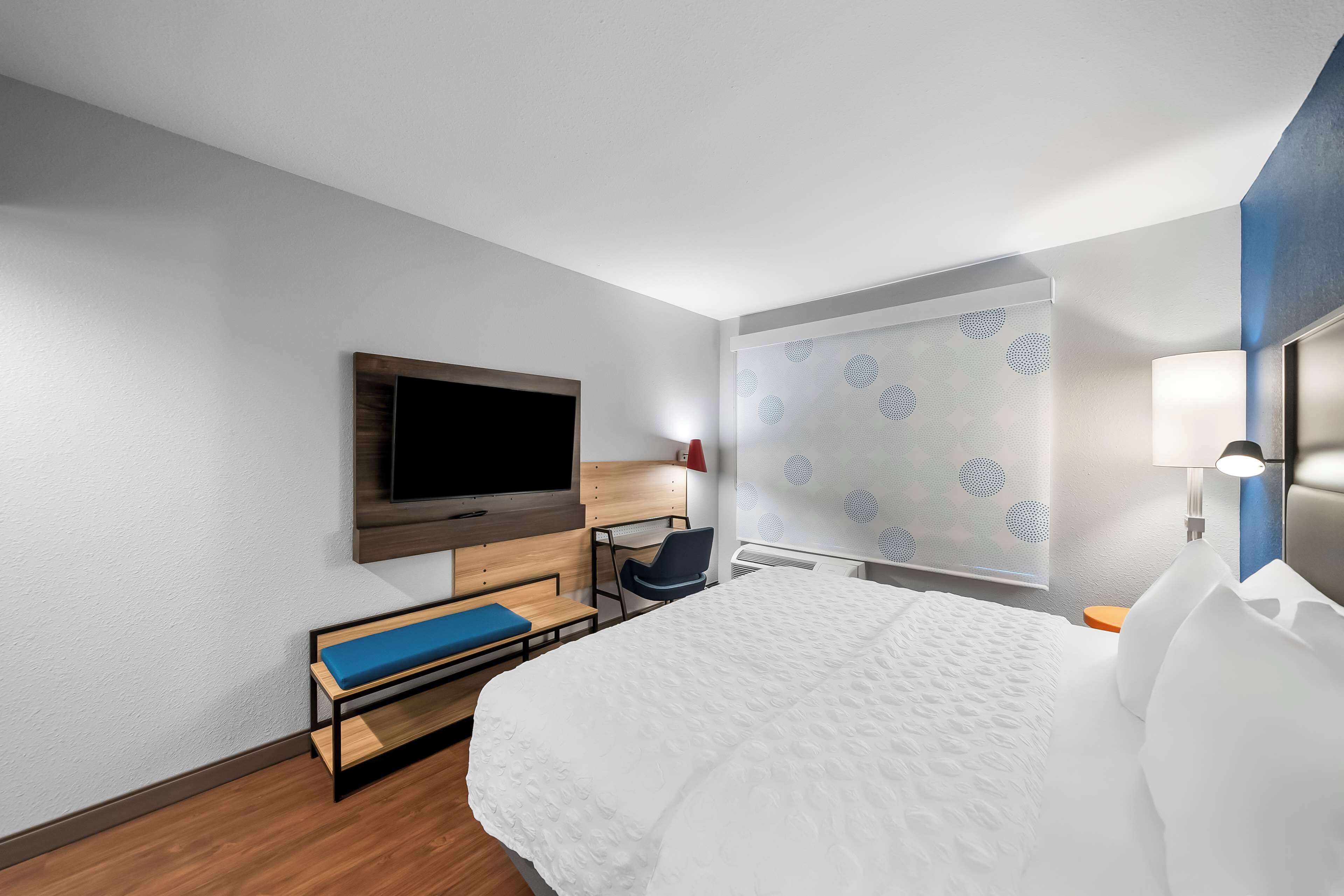 Habitació d'hotel amb llit, televisió penjada a la paret, escriptori amb cadira i finestra amb cortines estampades.