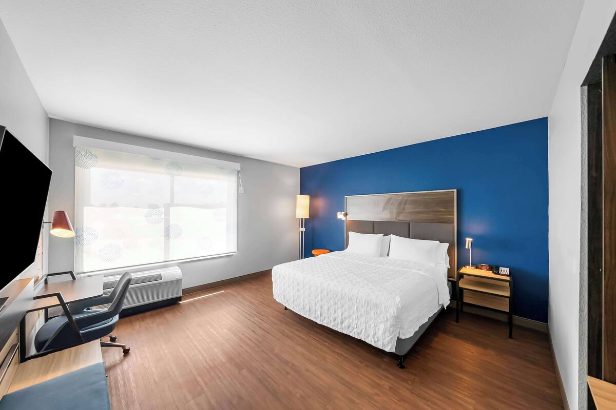 A modern hotel room with a large bed, a desk with a chair, a TV, and a window with blinds.
