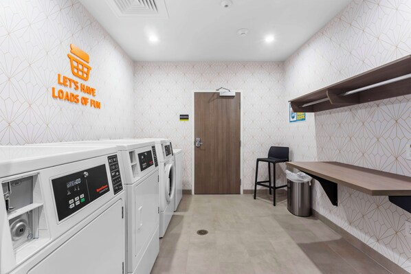 A laundry area with multiple washing machines, a wooden door, a trash bin, and a shelf. - Home2 Suites By Hilton Lake Jackson (Lake Jackson)