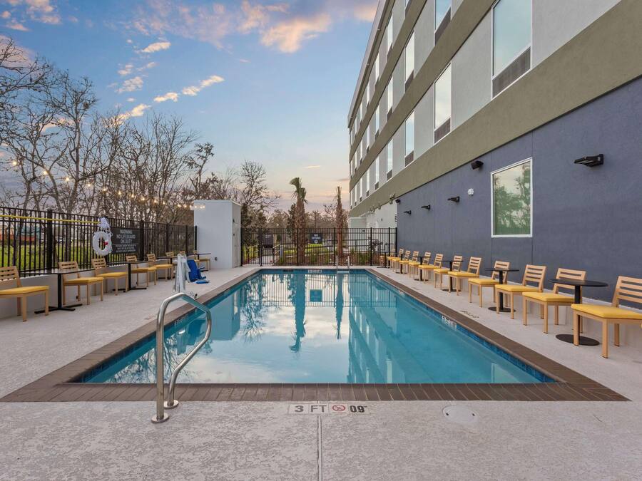 A swimming pool with a 3 FT depth marker, surrounded by yellow chairs and tables, with a building and trees in the background.