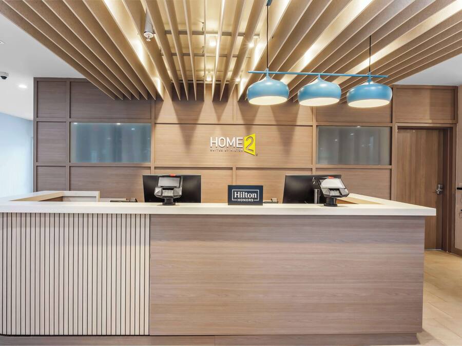 Un moderno mostrador de recepción de hotel con acabado de madera, un cartel que dice 'HOME 2' y dos lámparas colgantes azules arriba.