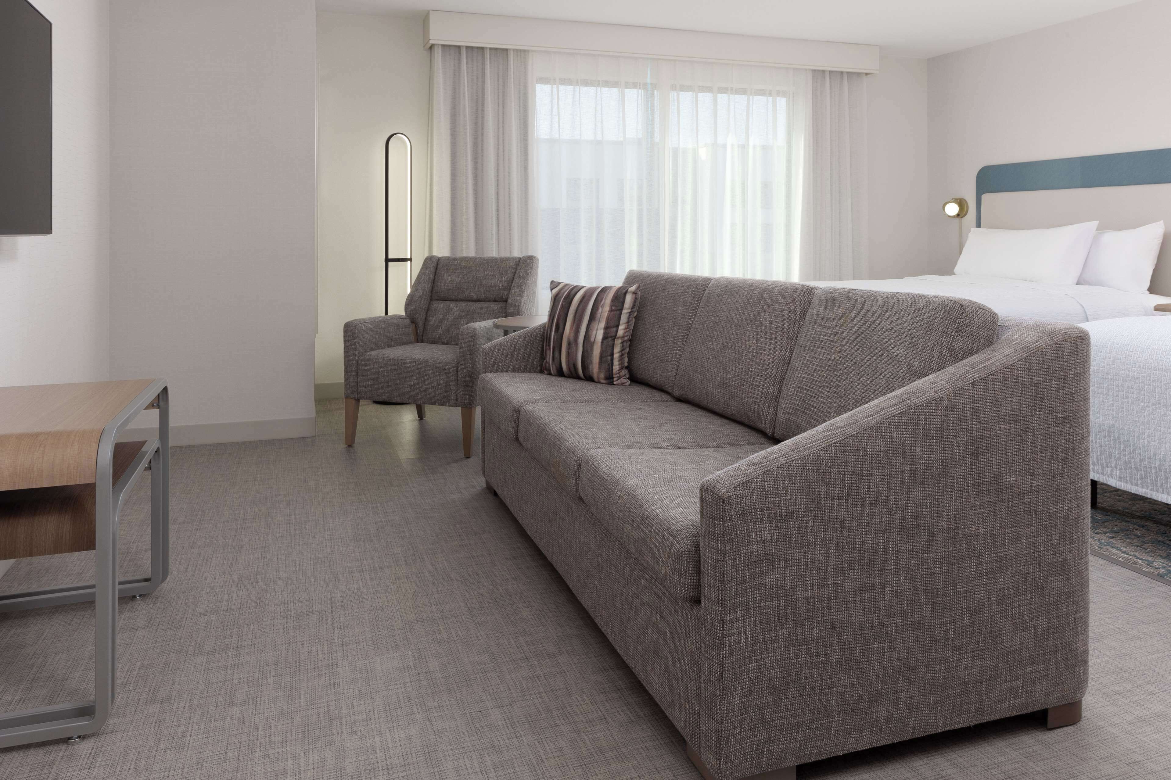 A hotel room with a grey sofa, a small armchair, a wooden side table, and a bed with white bedding.