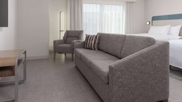 Una habitación de hotel con un sofá gris, un sillón pequeño, una mesita de madera y una cama con ropa blanca.