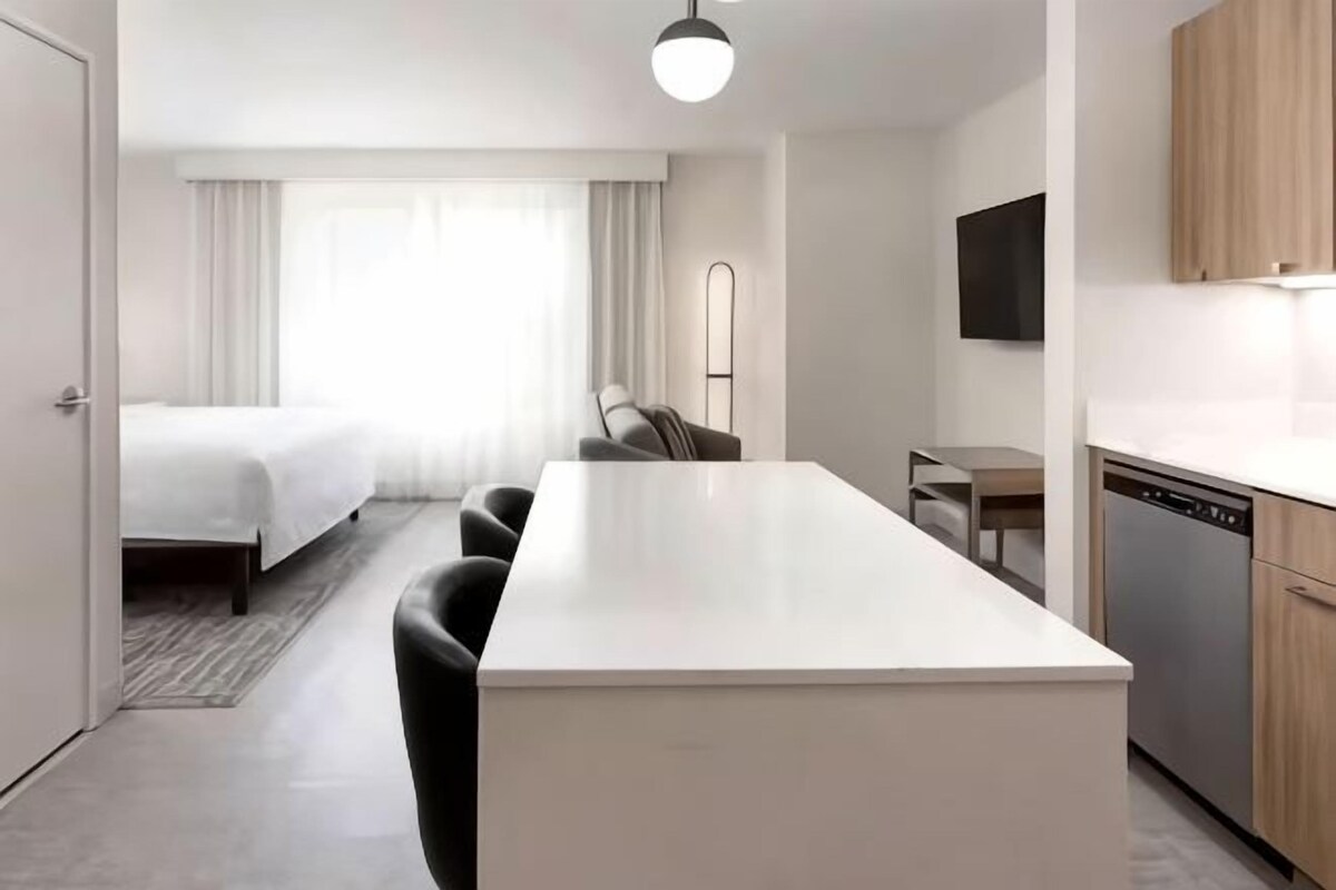 Una habitación de hotel moderna con una cama grande, una zona de comedor, una cocina con lavavajillas y un televisor montado en la pared.