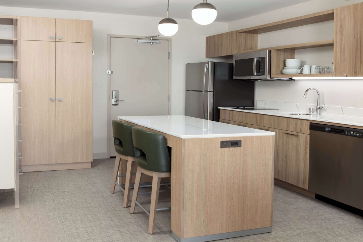 Eine moderne Küche mit Holzschränken, einem Edelstahlkühlschrank, einer Mikrowelle und einem Geschirrspüler. Eine kleine Kochinsel mit Stühlen und eine Tür mit Griff sind ebenfalls vorhanden.