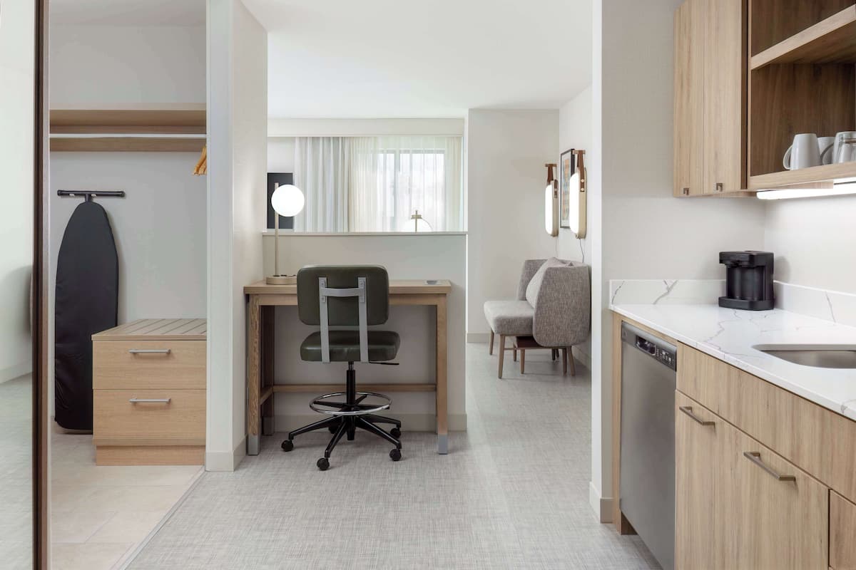 Ein modernes Hotelzimmer mit Schreibtisch aus Holz, einem Sessel, einem kleinen Esstisch, einer Couch, einer Kaffeemaschine und einem Geschirrspüler.