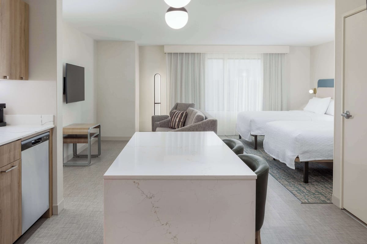 Ein modernes Hotelzimmer mit Marmortisch, einer Couch, zwei Einzelbetten, einem Flachbildfernseher und einem großen Fenster mit Vorhängen.