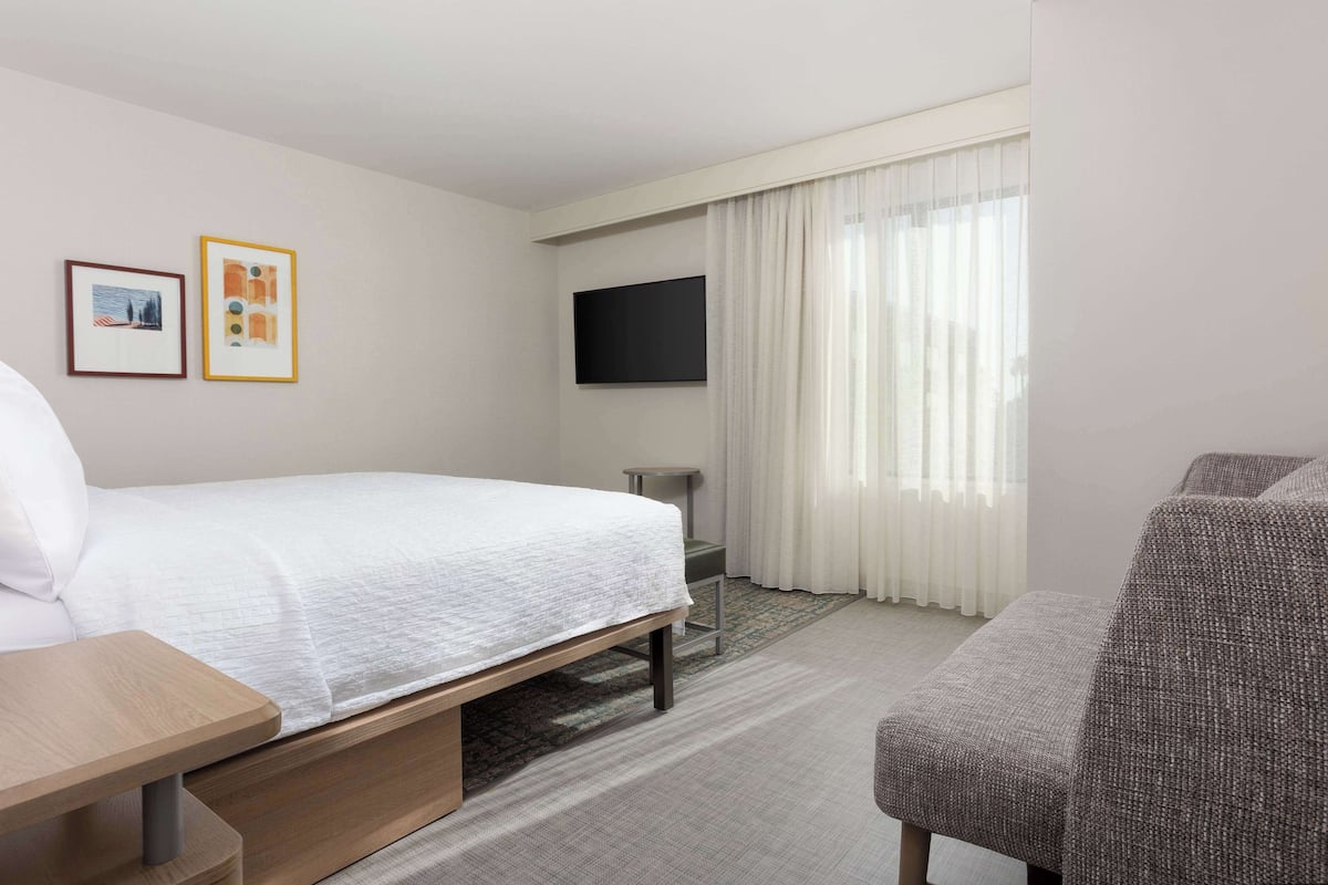 Ein Hotelzimmer mit Bett, Wandfernseher, Sessel und kleinem Tisch.