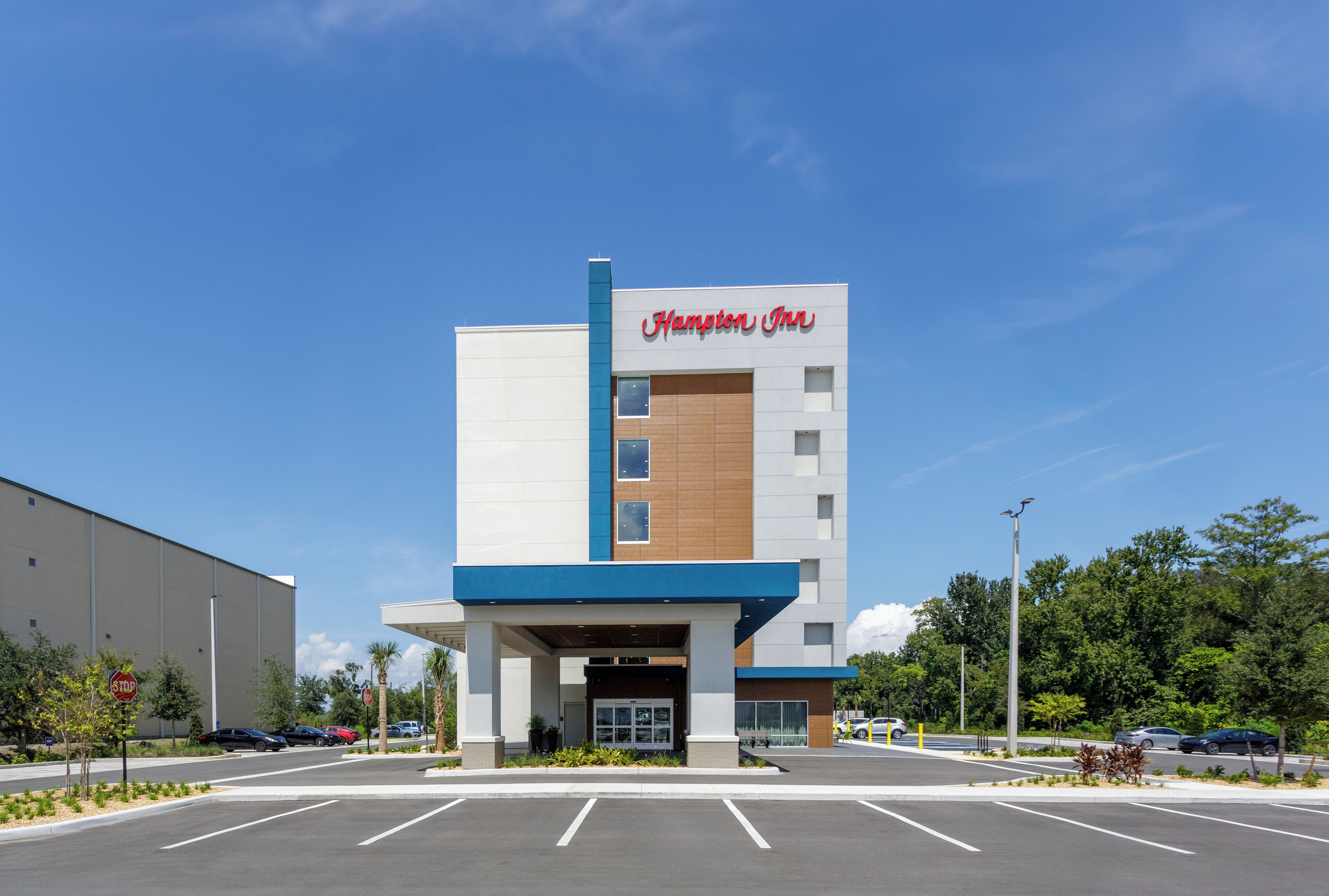 Un edificio moderno con el cartel de "Hampton Inn", un estacionamiento y un cielo azul despejado.