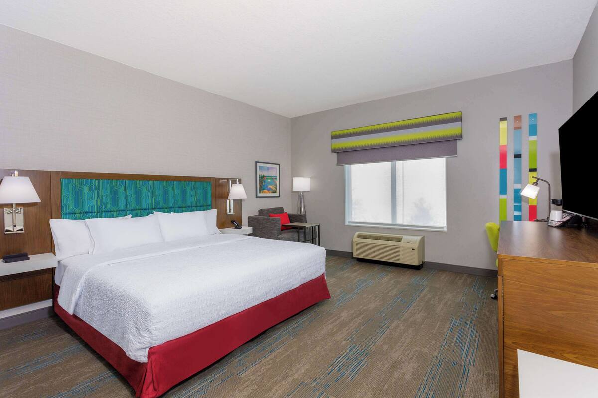 A hotel room with a large bed, a desk, a chair, a TV, a window, and a painting on the wall.