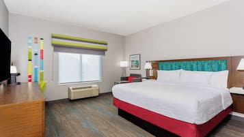 A hotel room with a large bed, a desk, a chair, a window with blinds, a wall-mounted air conditioner, and a framed picture on the wall.