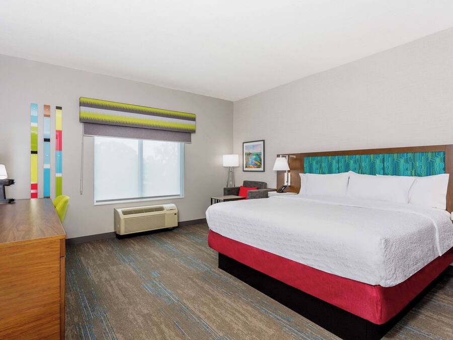 A hotel room with a large bed, a desk, a chair, a window with blinds, a wall-mounted air conditioner, and a framed picture on the wall.