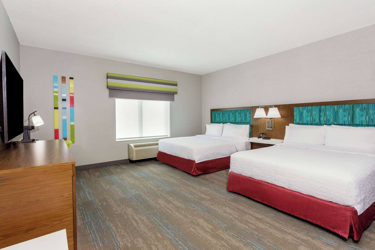 A hotel room with two beds, a desk, a TV, and a window with blinds.